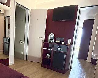 Interno di una camera d’albergo con TV, minibar, cassaforte e porte aperte.